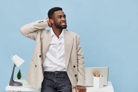 Professional man in casual business attire poses in contemporary office environment with a light blue backdrop, expressing thoughtfulness or surprise, standing next to a desk with a computer, lamp, and stationery, conveying modern work atmosphere and confidence. Concept: workplace professionalism
