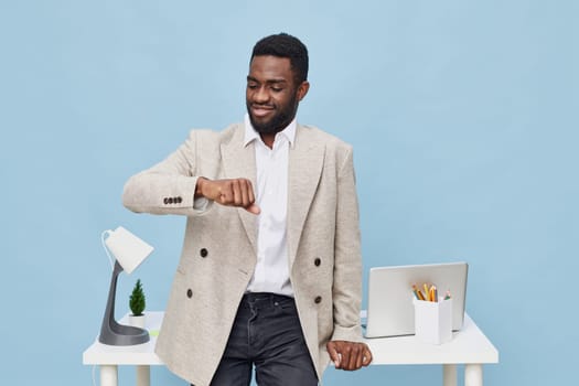 Professional young man of African descent in beige blazer and white shirt looking at his watch in bright office environment with desk, laptop, stationery, and pastel blue backdrop. Concept: time awareness and productivity.