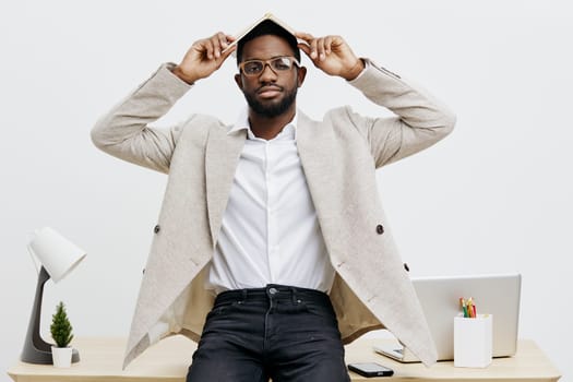 Confident African American man with glasses and a beard, sitting at a sleek desk, adjusting his hat, dressed in a beige blazer and white shirt. Modern office background with plant, lamp, and stationery, emphasizing a professional and stylish work atmosphere. Concept: success and style.