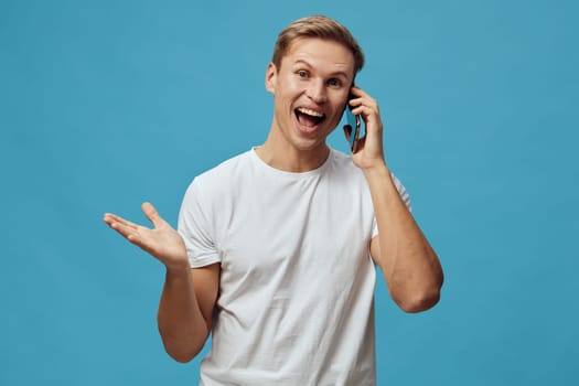 Happy man engaging in phone call on a smartphone, wearing a casual white t-shirt, smiling widely, friendly mood, plain blue background, studio setup, informal communication, positive emotion, technology use. Concept: communication enjoyment