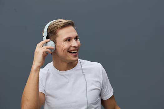 Happy young man with short brown hair, dressed casually in a white t-shirt, enjoying music through wireless headphones, smiling and looking to the side against a plain gray backdrop, studio shot emphasizing leisure and entertainment. Concept: joy of music
