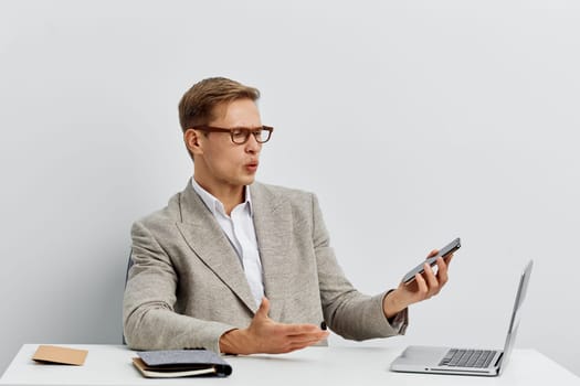 surprised man using smartphone and laptop for digital nomad remote work with glasses and casual jacket on plain white background. He communicates online multitasking with technology gadgets. Concept: digital nomad