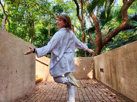 Mature blonde woman playfully spinning in a lush green environment, embracing the joy of travel and adventure on her exciting trip.