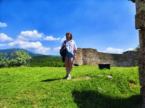 A woman enjoys her adventure, standing near historic ruins under a clear blue sky surrounded by lush greenery.
