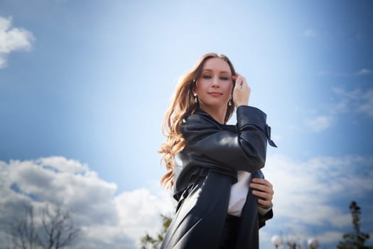 A young woman stands outdoors under a cloudy sky, showcasing her stylish outfit and natural beauty in sunlight.