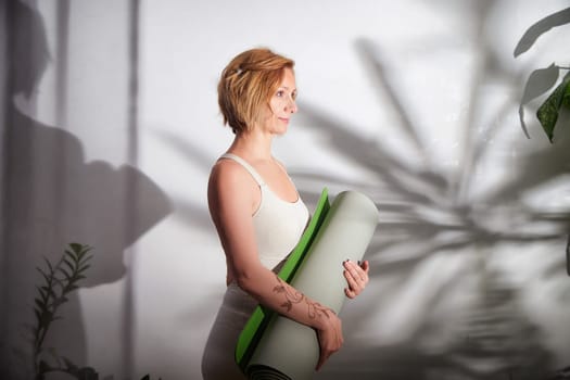 A woman stands still, holding a yoga mat, embracing a moment of calm in a peaceful indoor setting with natural light.