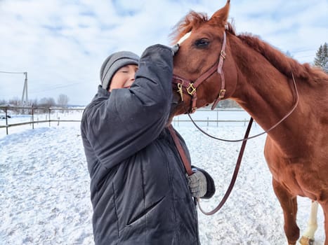 A person warmly engages with a horse during winter, enjoying the snowy landscape and bright, cloudy skies.