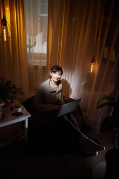 In a warm and inviting apartment, a middle-aged woman relaxes in the evening light, focused on her laptop while sipping a drink.