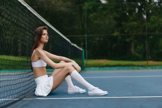 Confident young woman in white sportswear relaxing on a tennis court, showcasing her athleticism and determination, with vibrant green background adding freshness