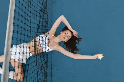 Joyful young woman dressed in sporty white apparel holding a tennis ball while lying on a blue court, embodying a healthy, active lifestyle in a vibrant setting