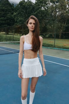 Confident young female tennis player in a white skirt and sports bra, showcasing athleticism on a court surrounded by lush greenery