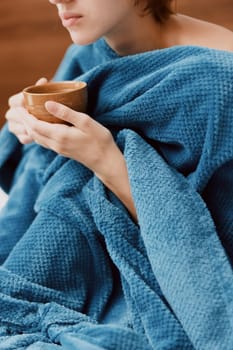 Woman wrapped in blue blanket holding cup of tea and wooden spoon.