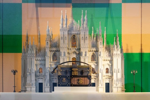 Milan, Italy - April, 08, 2025: Luxury Louis Vuitton handbag displayed in front of detailed Milan Duomo replica. Designer window display with colorful checkered backdrop.