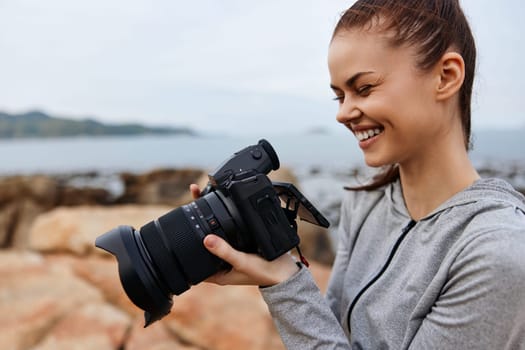 A cheerful young woman dressed in a gray hoodie, engaged in photography by the beach, embodying creativity, happiness, and a love for capturing moments