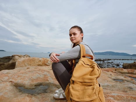 A young woman with a yellow backpack is crouching on rocky terrain by the sea, dressed in casual clothing, showcasing a reflective and thoughtful mood in a serene coastal environment