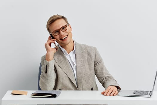 Confident smiling man with glasses in smart casual attire, sitting at a desk with a laptop and notepad, engaging in a phone call, indoor office setting, professional work atmosphere, modern business environment, success and communication concept