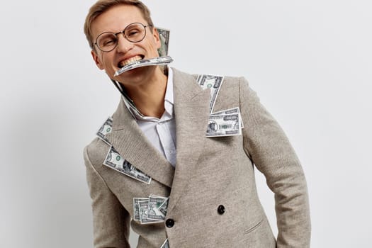 Cheerful young man with glasses and a big smile, displaying a bundle of cash in his mouth while dressed in a fashionable beige suit decorated with dollar bills, standing against a white studio background. Concept: financial success, humor, confidence