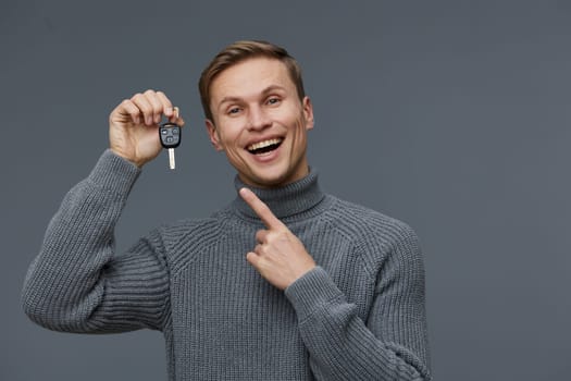 smiling young man holding car key wearing gray knitted sweater excited pointing isolated on gray background. Cybersecurity and digital nomad concept with casual autumn outfit and cheerful emotion.