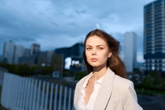 A poised business woman stands confidently in an urban environment, wearing a light-colored suit. The twilight backdrop creates a dynamic atmosphere, emphasizing her professional demeanor.