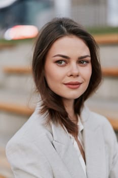 confident woman in beige blazer with subtle smile sitting outdoors with blurred urban background, natural light, neutral tones, fashion concept