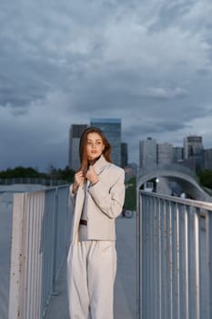 A confident woman in light beige suit stands on a metal urban bridge with skyscrapers and cloudy sky in the background, embodying modern business fashion and autumn mood.
