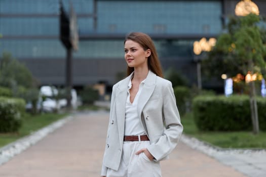 A professional businesswoman in a light-colored blazer and trousers confidently strolls through an urban landscape, exuding poise and sophistication in her demeanor.
