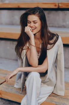 Young woman in beige business suit smiles confidently while sitting on wooden steps outdoors in bright natural light, showcasing casual fashion and positive emotion in spring.