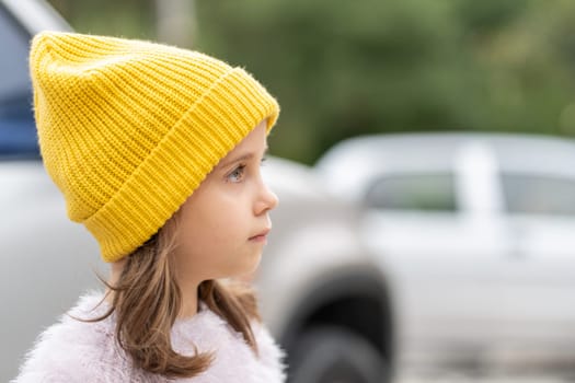 Side view of young girl wearing bright yellow knit beanie outdoors. Candid portrait captures thoughtful expression on child's face. Blurred background with cars visible
