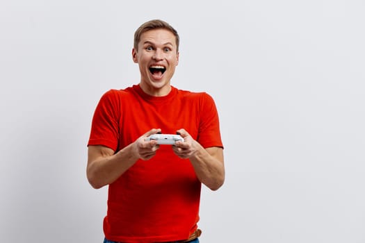 Happy cheerful man in casual red t-shirt enjoying gaming session with game controller, showing excitement and enthusiasm, against a simple white background. Concept: leisure and entertainment.
