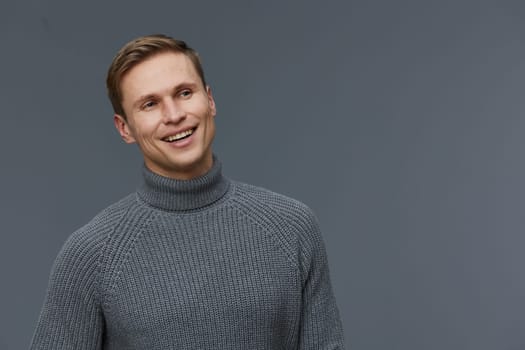 Happy young man wearing a gray turtleneck sweater and smiling brightly while standing against a neutral gray backdrop in a studio setting. The image captures a cheerful, relaxed mood, highlighting modern casual fashion and positive emotions. Concept: joy of casual style