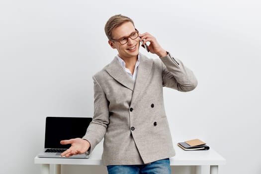 smiling man talking phone in casual jacket and jeans standing near desk with laptop and notebook, expressing positive communication and remote work lifestyle, digital nomad concept involving videoconference and flexible job environment