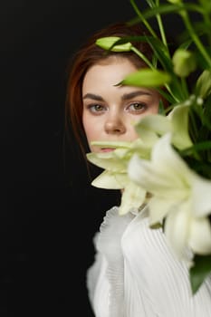 A young model with soft natural makeup, dressed in white, presents a serene expression with flowers, set against a dark background, ideal for beauty and lifestyle concepts