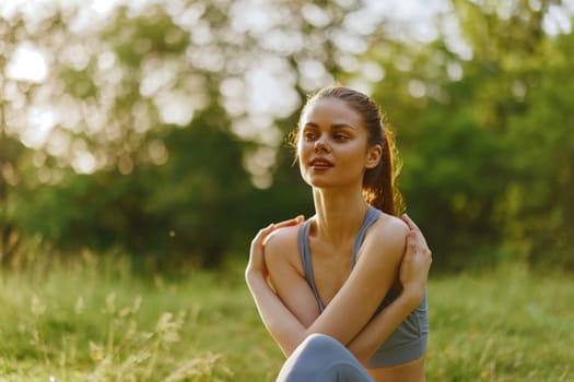 Athletic young woman in sports attire peacefully sitting in a sunlit park, embracing nature and conveying relaxation amidst lush greenery, ideal for wellness themes