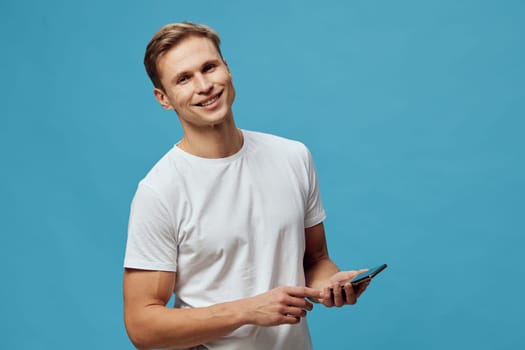 Happy young man with short hair dressed in casual white t-shirt holding a smartphone while looking at camera against a vibrant blue background. Concept: technology, communication, youth, positivity, digital lifestyle