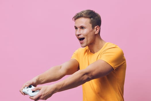 Smiling enthusiastic man having fun gaming, holding game controller with both hands, expressing joy and excitement, dressed in casual orange shirt, pink studio background, lifestyle, leisure, technology concept