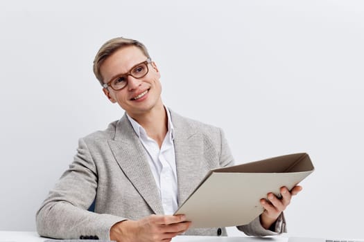 Smiling businesswoman with short hair and glasses, dressed in a light gray blazer and white shirt, holding a folder in her hands. Bright office environment, positive mood, professional appearance, corporate attire, success concept, modern work setting.