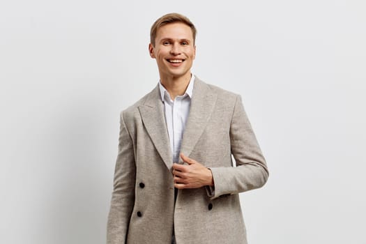 Young man with cheerful smile wearing elegant light gray suit and white shirt, posing confidently with hand on chest, plain white studio background, professional business attire, positive mood, fashion style, indoor photography, lifestyle concept