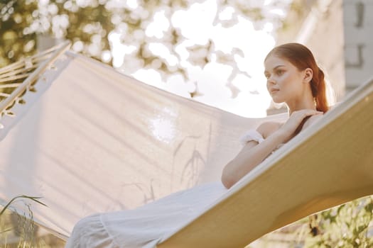 A peaceful young woman in a flowing white dress lounges on a hammock, basking in soft sunlight and surrounded by greenery, evoking a sense of serenity and relaxation