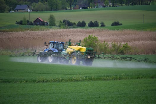 Farm tractor with sprayer disperses liquid over a lush green field amid a picturesque rural countryside setting. Surrounding the scene are farmhouse buildings, trees and vibrant agricultural landscape