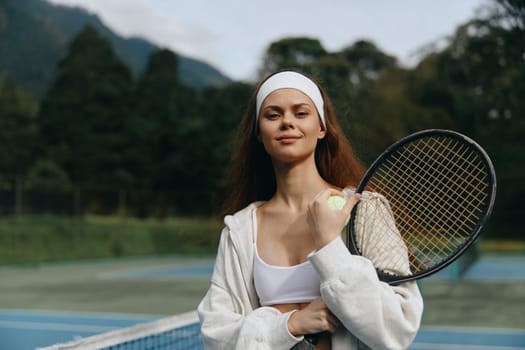 Confident young female tennis player in a sporty outfit and headband, smiling on the court with a picturesque nature background, embodying sportsmanship and fitness