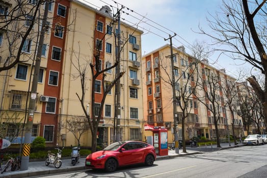 Shanghai, China - Quiet residential street in Pudong with parked scooters, red Tesla and vintage telephone booth. Yellow-terracotta apartments, bare tree, urban atmosphere