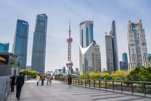 Shanghai, China - 1 April 2025: Futuristic Pudong district with pedestrians on an overpass. Oriental Pearl Tower and glass skyscrapers under blue sky
