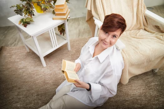 A middle-aged woman relaxes in a cozy room, reading a book while seated on a soft rug surrounded by books.
