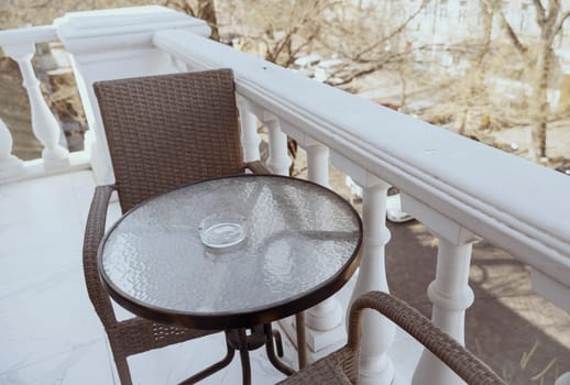 On the balcony there is a table, an armchair, an ashtray on the table. A glass table with a crystal, clean ashtray on it, next to it a brown wicker armchair. A balcony in a classic style