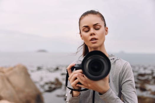 A young woman photographer in a hoodie, focused on her camera by the ocean The cloudy sky and rocky background enhance the creative atmosphere, ideal for photography enthusiasts