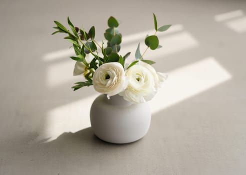 White ranunculus flowers paired with green foliage are creatively arranged in a minimalist vase. Soft natural light enhances the serene atmosphere of the setting.