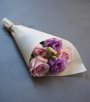 A bouquet featuring pink roses and purple flowers is elegantly wrapped in simple paper. This floral arrangement is placed on a smooth gray surface, highlighting its delicate colors.