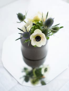 A delicate arrangement of white flowers and thistles sits in a simple vase on a smooth, reflective table surface, illuminated by gentle natural light.