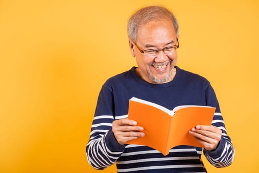 Portrait smiling Asian old man wearing glasses reading a book studio shot isolated yellow background, happy elderly man enjoying interesting story, time to relaxation