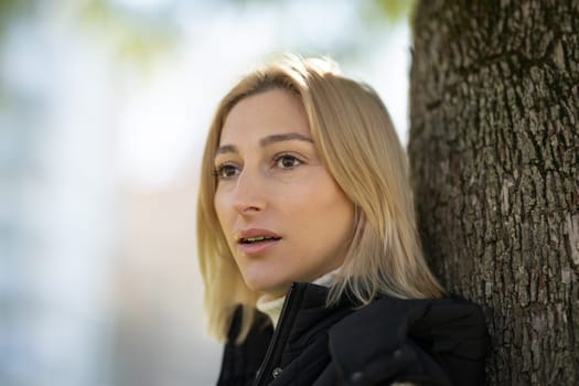 Blond woman with turtleneck sweater and black jacket leaning against tree looking away in thought